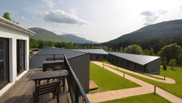 A modern building with a deck and outdoor seating area, surrounded by a well-maintained lawn and a mountain backdrop. - Bridge of Orchy Hotel (Bridge of Orchy)
