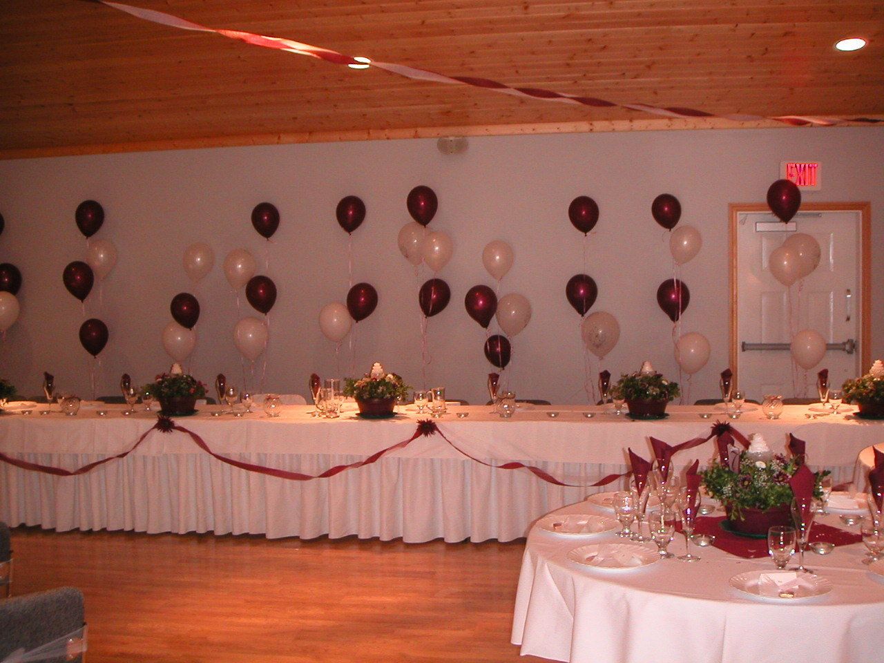 A banquet hall with tables set for a formal event, featuring white tablecloths, red ribbons, and floral centerpieces.