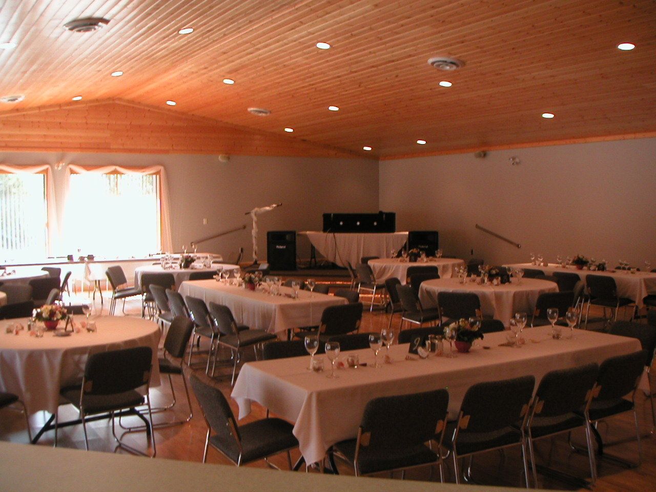 A banquet hall with multiple round tables, each set with chairs and table settings, featuring a wooden ceiling and large windows.