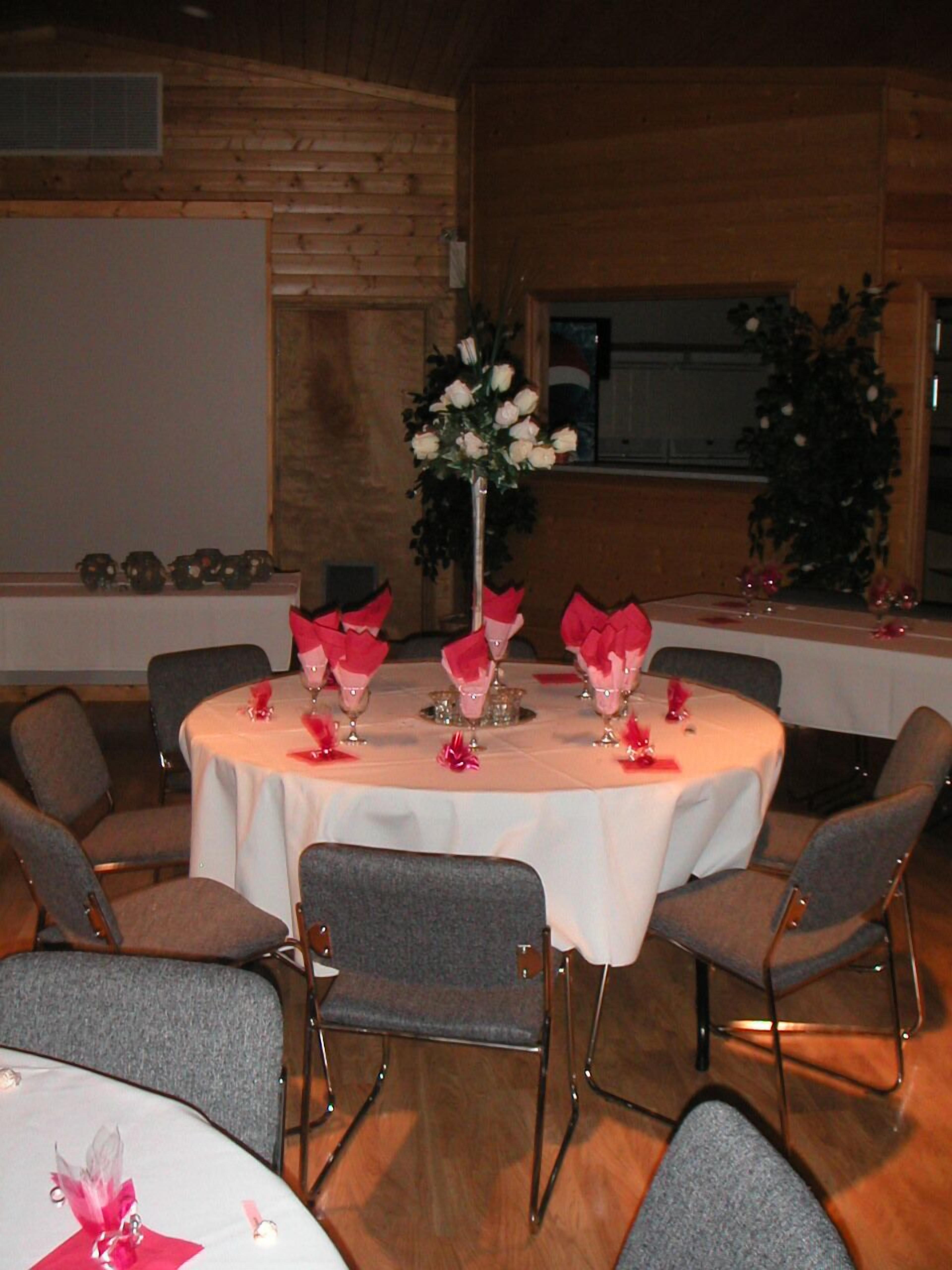 A round table with a white tablecloth, surrounded by grey chairs, in a room with wooden walls and a window.