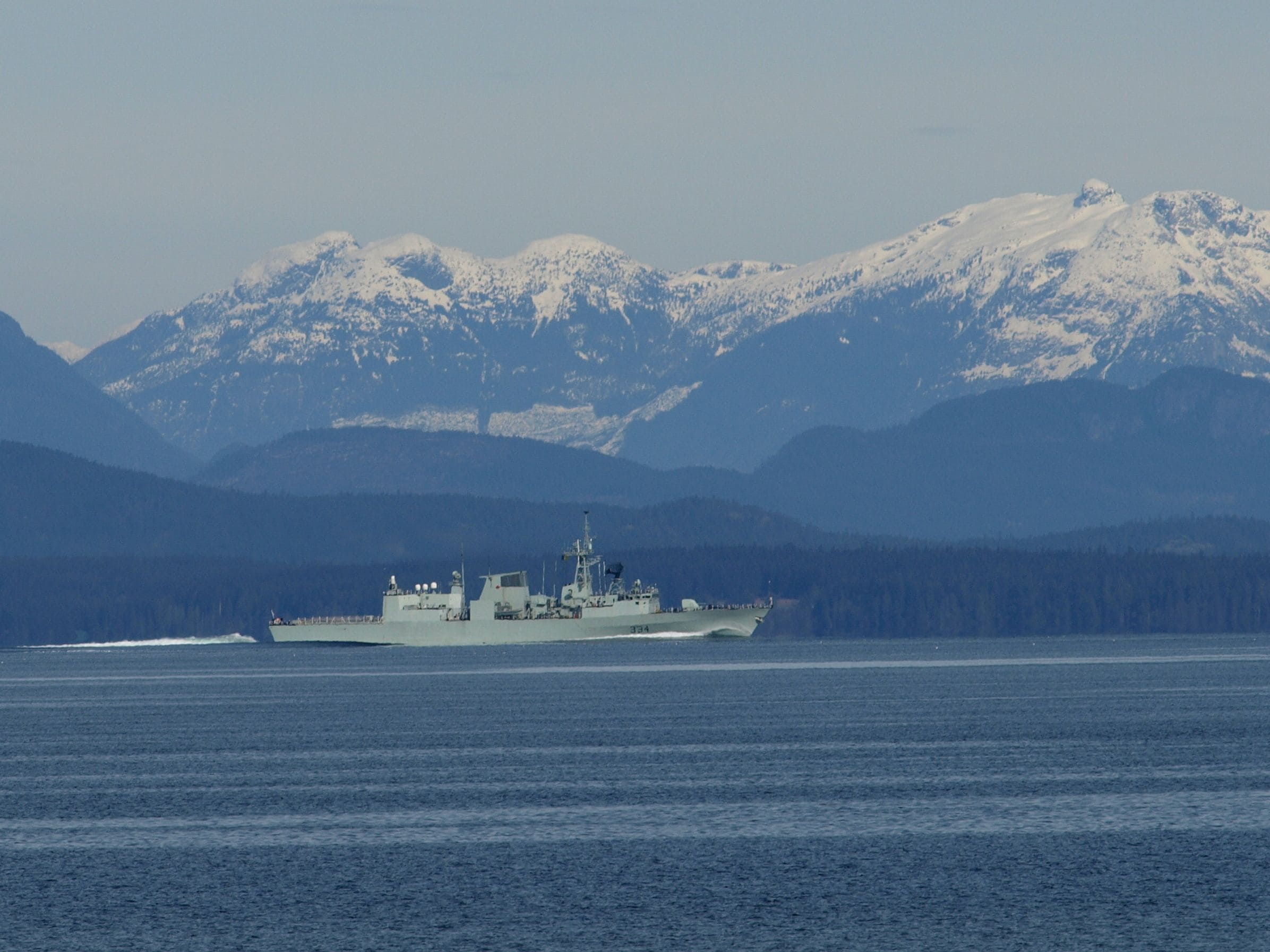 A military ship on calm waters with snow-capped mountains in the background.