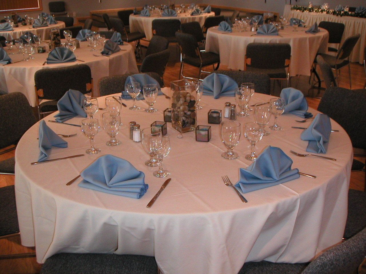 A banquet hall with round tables set for dining, each with a white tablecloth, blue napkins, and silverware.