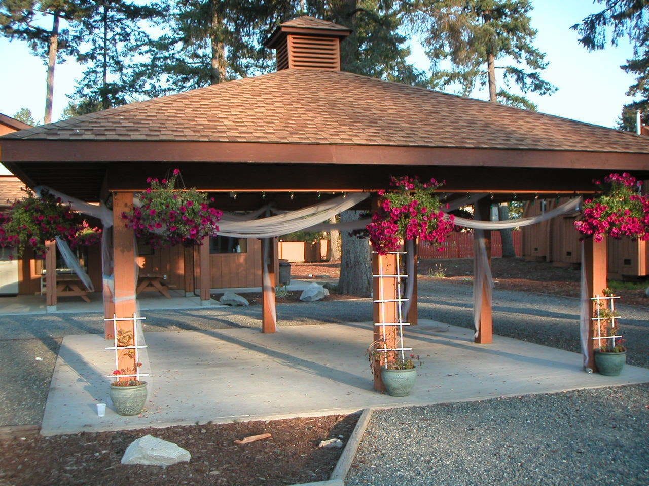 A gazebo with a tiled roof, supported by wooden pillars, and surrounded by potted plants with pink flowers.