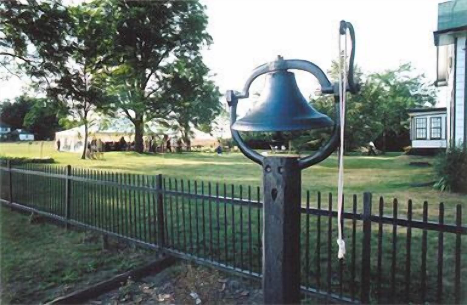 A black metal bell mounted on a post with a curved top, located in a grassy area with a metal fence in the foreground.