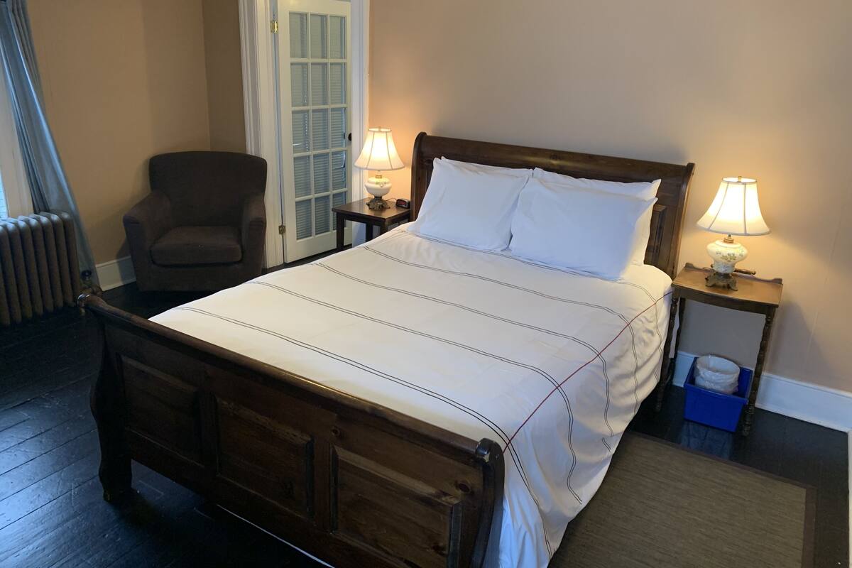 Una cama bien hecha con cabecera de madera, dos lámparas de noche, una silla y una puerta con paneles de vidrio.