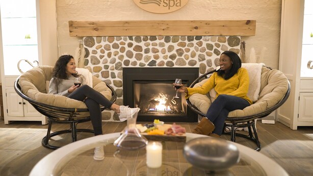 Two people seated in cushioned chairs facing a fireplace, with a glass table in front of them. - Stevenson Inn & Spa (Essa)
