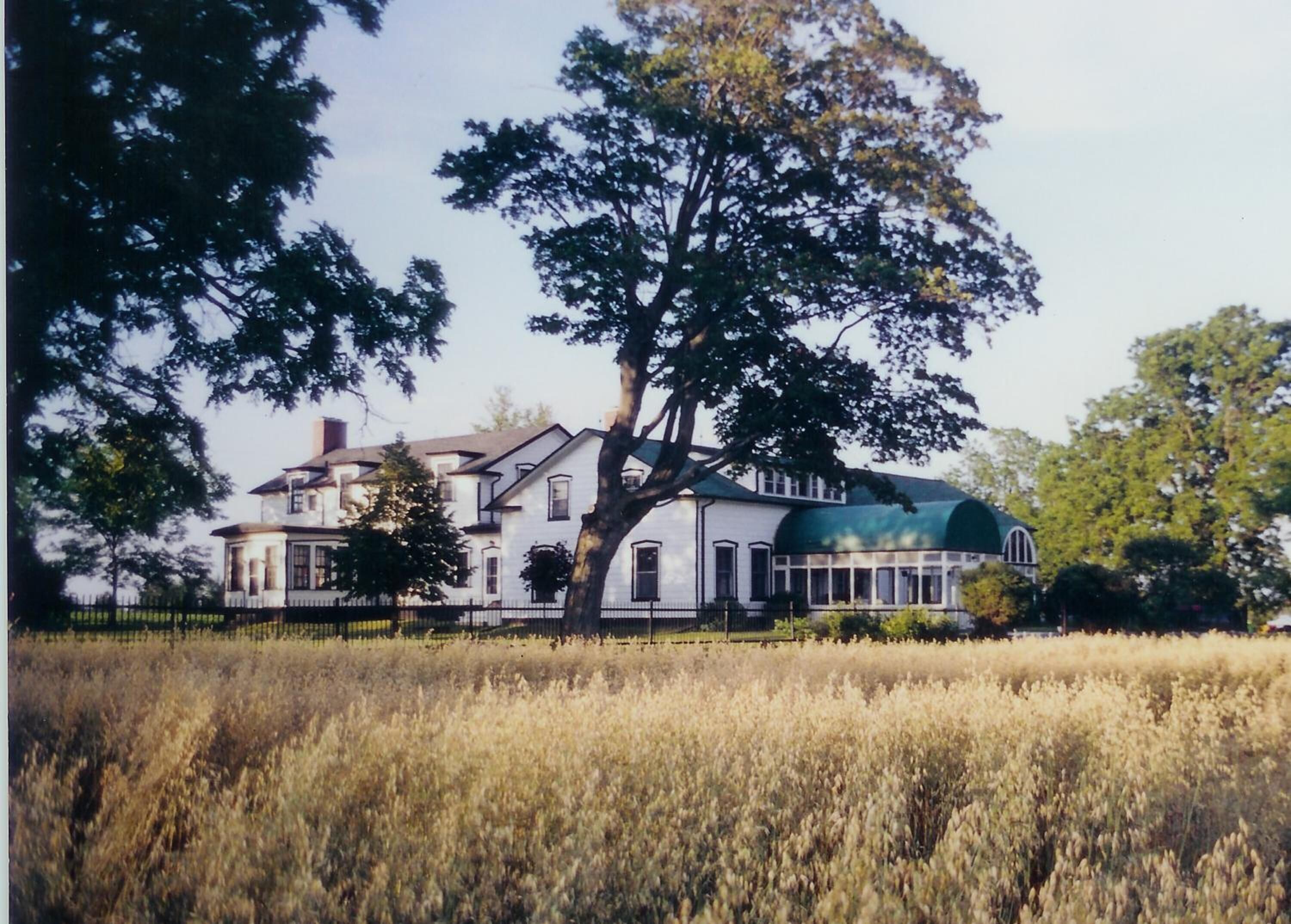 Een groot, twee verdiepingen tellend huis met een groen dak en veel ramen, omringd door een veld en bomen.