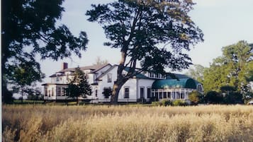 A large, two-story house with a green roof and multiple windows, surrounded by a field and trees.