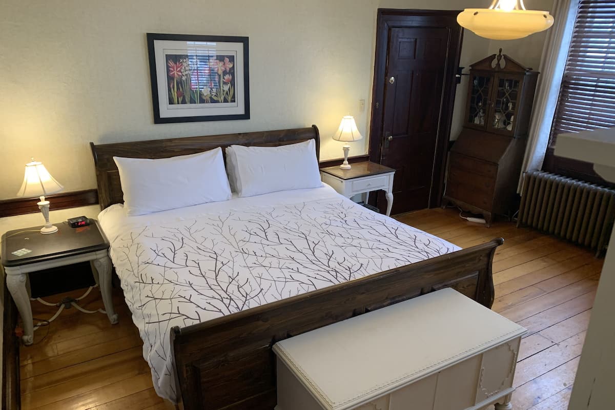 Un dormitorio con cama de madera, ropa de cama blanca, mesita de noche con lámpara, cómoda y un cuadro en la pared.