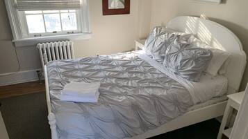 Een net opgemaakt bed met een grijs gebonden beddekleed en kussens, een wit hoofdbord, een nachtkastje en een radiator.