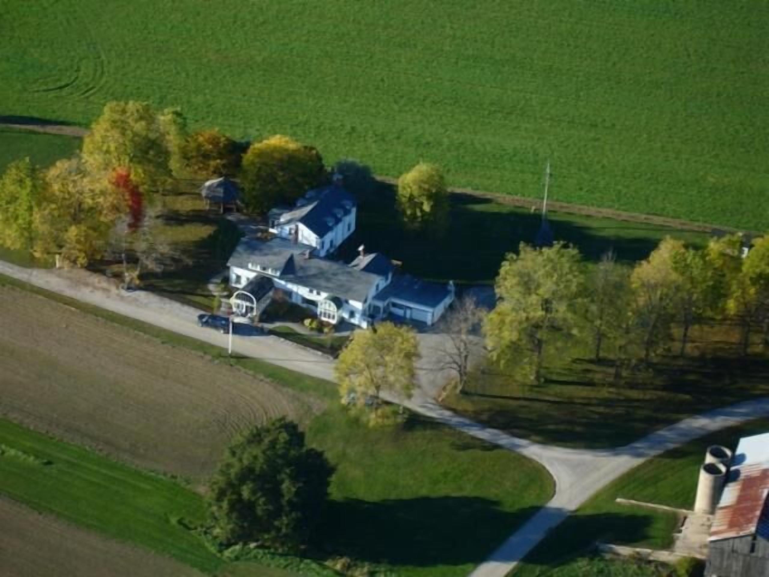 Eine Vogelperspektive eines großen, zweistöckigen Hauses mit markantem Schornstein, umgeben von grünen Feldern und Bäumen.