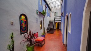 Un estrecho pasillo con suelo de baldosas rojas, paredes azules y una escalera que lleva a un nivel superior. Se observan plantas en macetas y un banco a lo largo de las paredes.