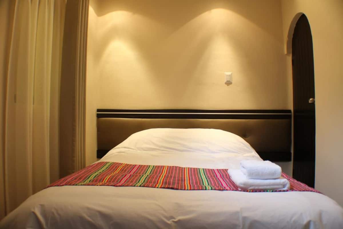 A hotel room with a bed, a headboard, a wall-mounted light, and a door.