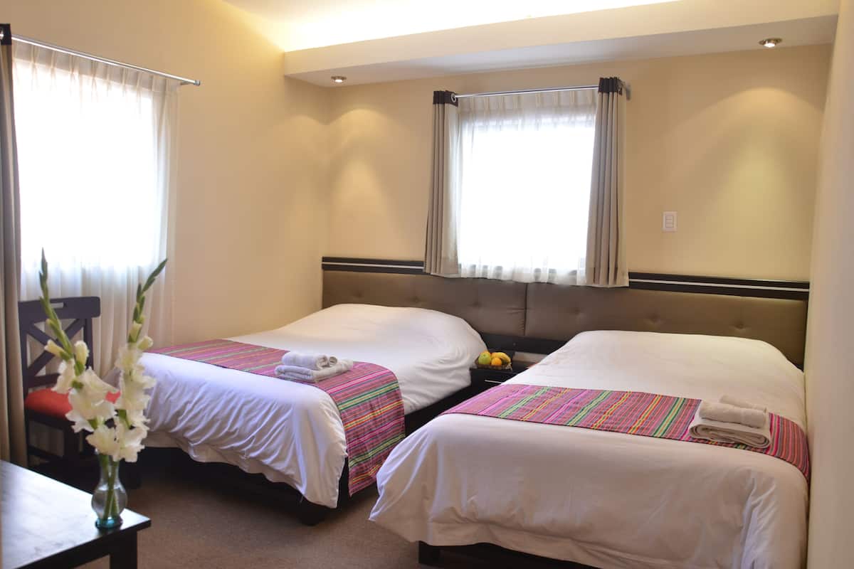 A hotel room with two beds, each with white linens and striped bedspreads. There is a vase with flowers on a table, and a window with curtains.
