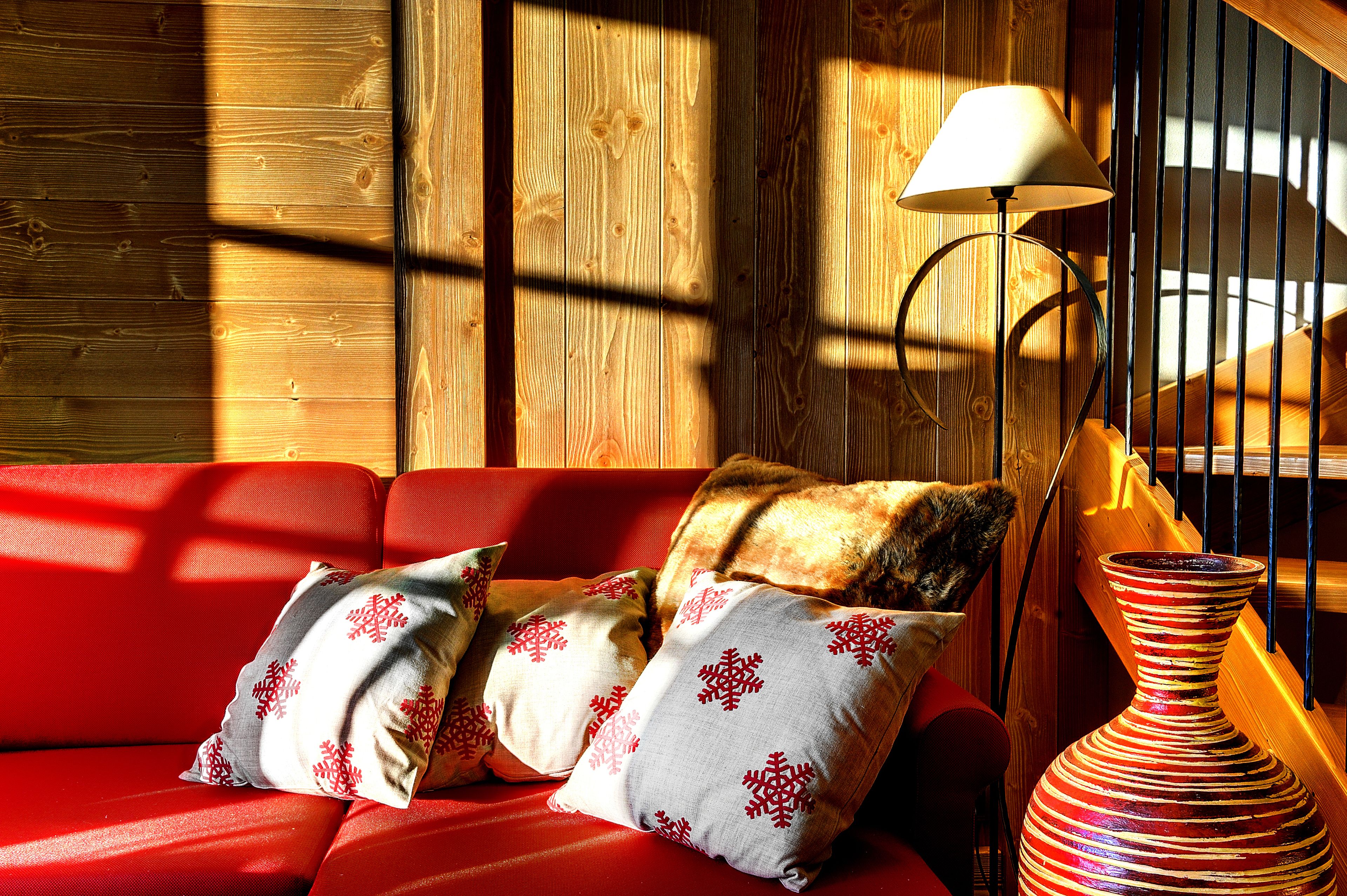 Un intérieur en bois avec un canapé rouge, des coussins à motifs, un lampadaire et un vase.