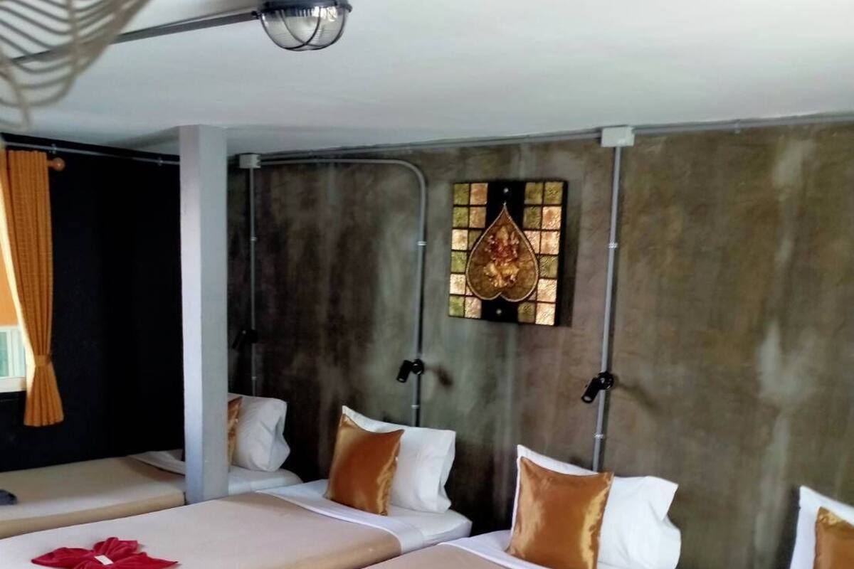 A hotel room with four single beds, each with a neatly folded towel. The room has a modern design with a large window, a wall-mounted light fixture, and a decorative glass panel.
