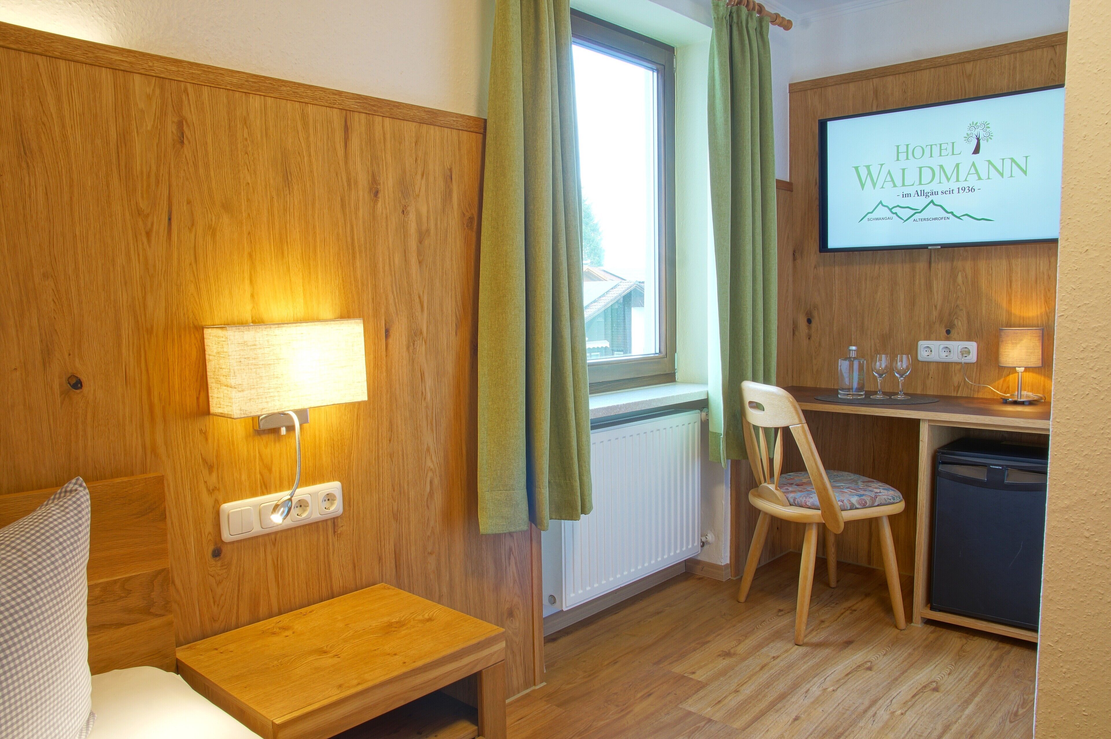 A hotel room with a wooden wall, a bed, a desk with a chair, a small table, a radiator, and a TV displaying the hotel name.