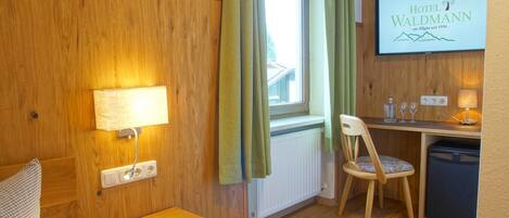 A hotel room with a wooden wall, a bed, a desk with a chair, a small table, a radiator, and a TV displaying the hotel name.