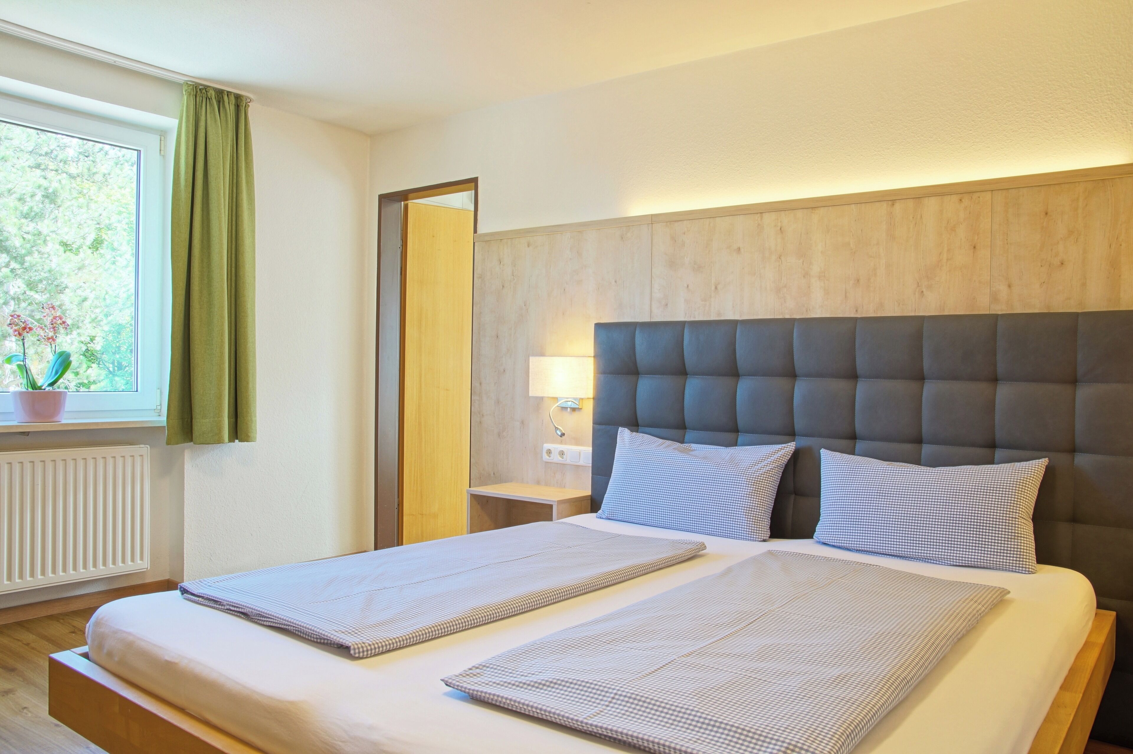 Een netjes opgemaakt bed met hoofdbord, een nachtkastje met lamp en een raam met gordijnen.