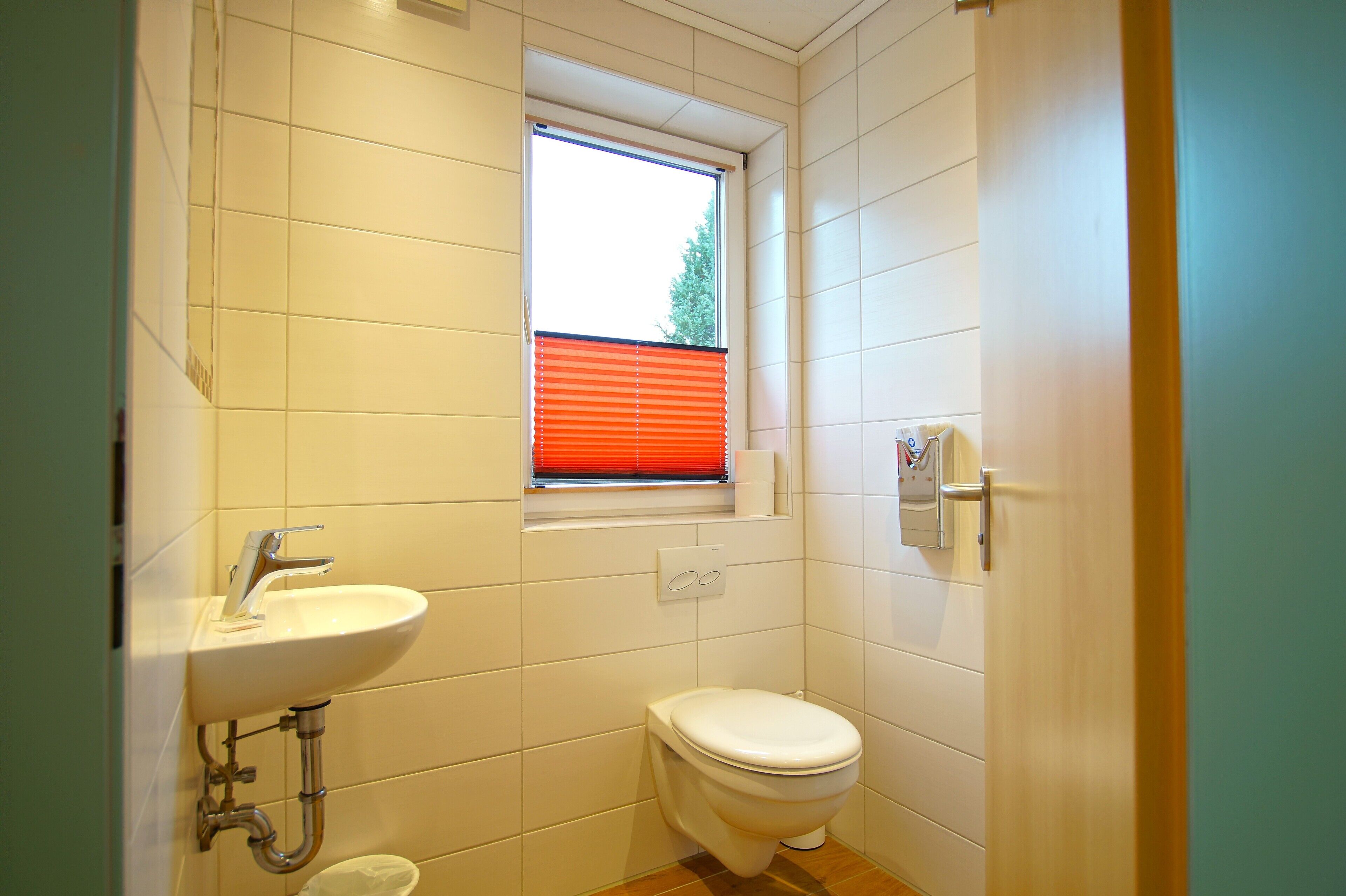 Een kleine badkamer met een witte wastafel, toilet en raam met rode lamellen.
