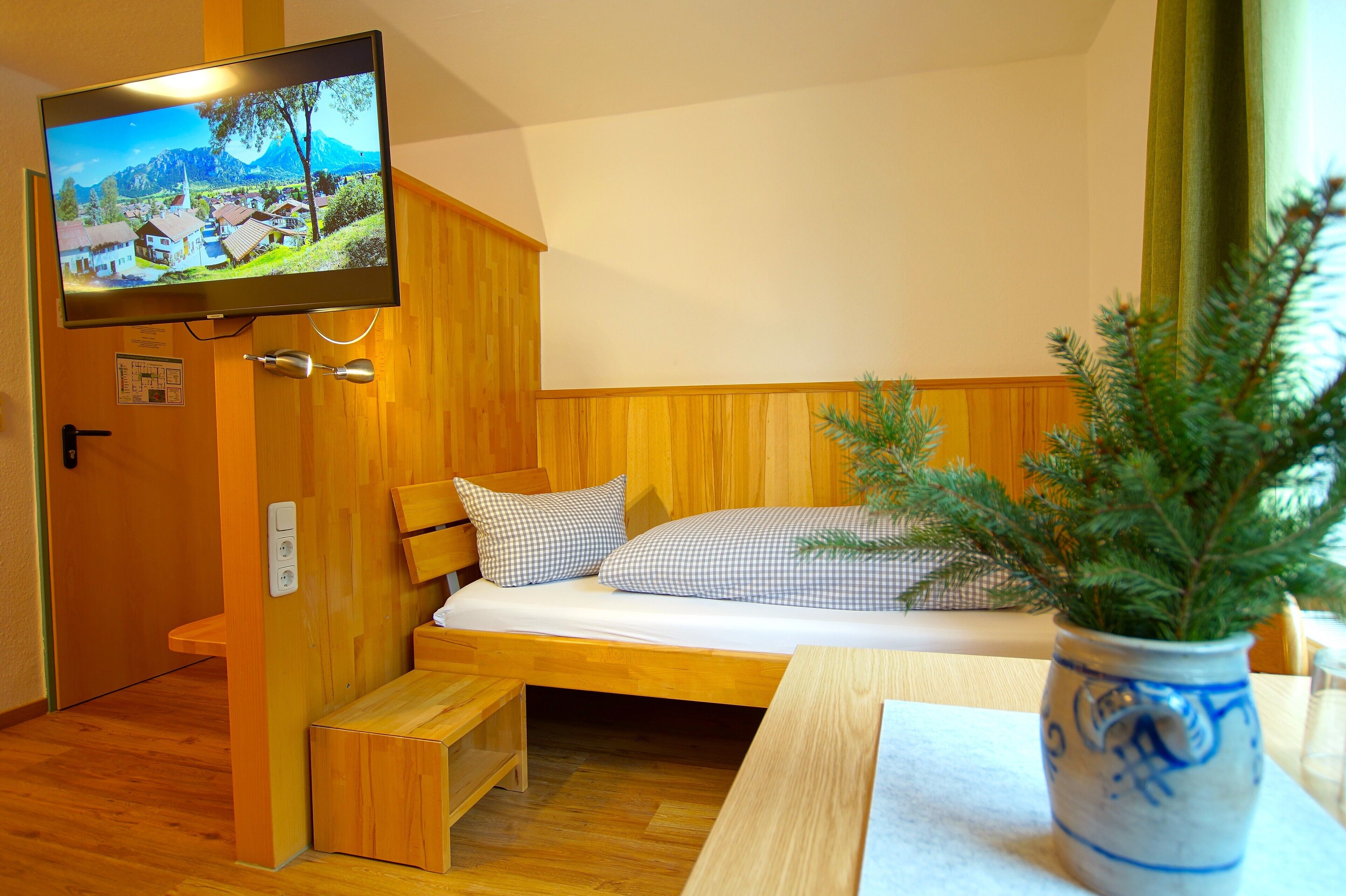 Habitación compacta con una cama individual, una mesita pequeña, un televisor montado en la pared y un armario de madera.