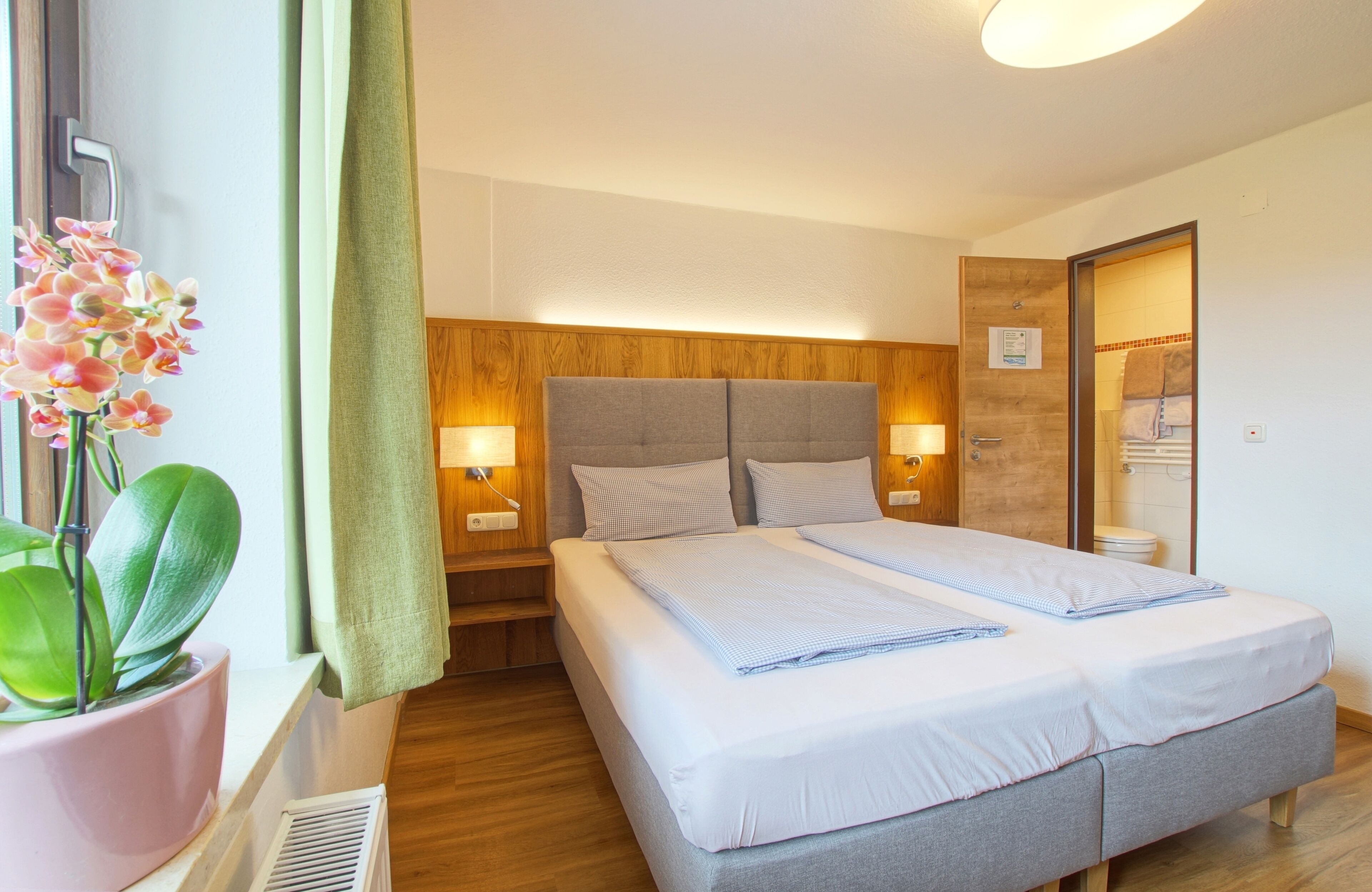 A hotel room with a double bed, wooden headboard, bedside lamps, a potted orchid, and a window with curtains.