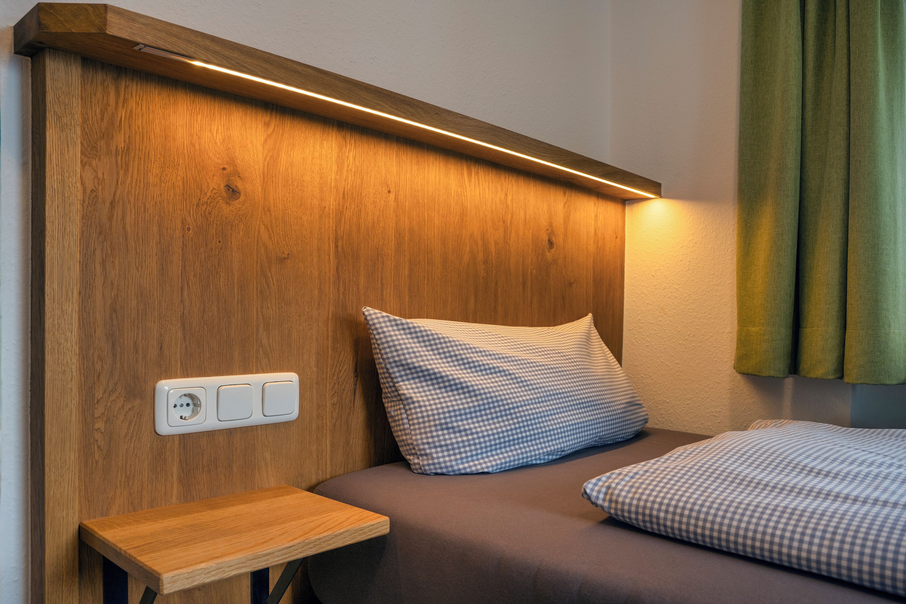 Ein hölzerner Kopfseitenverzierer mit integrierter Beleuchtung, ein Nachttisch und ein Bett mit Karomuster-Bettwäsche.