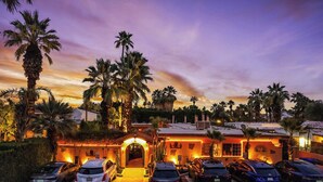 A hotel with a driveway leading to a gated entrance, surrounded by palm trees and a sunset sky. - Coyote Inn - 21 and Over - A Town & Desert Boutique Hotel (Palm Springs)