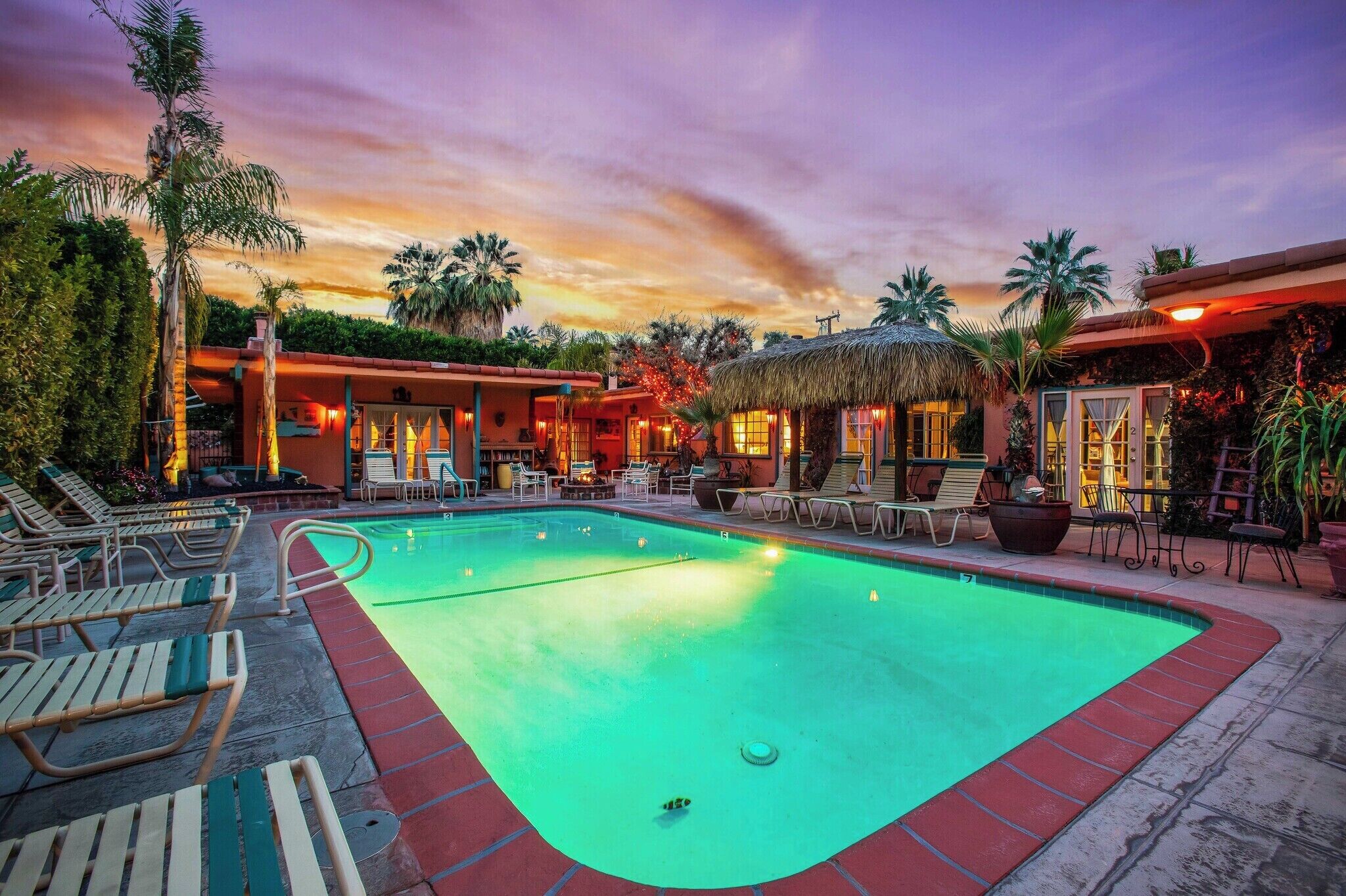 A swimming pool with lounge chairs and a thatched-roof structure, surrounded by palm trees and a sunset sky.