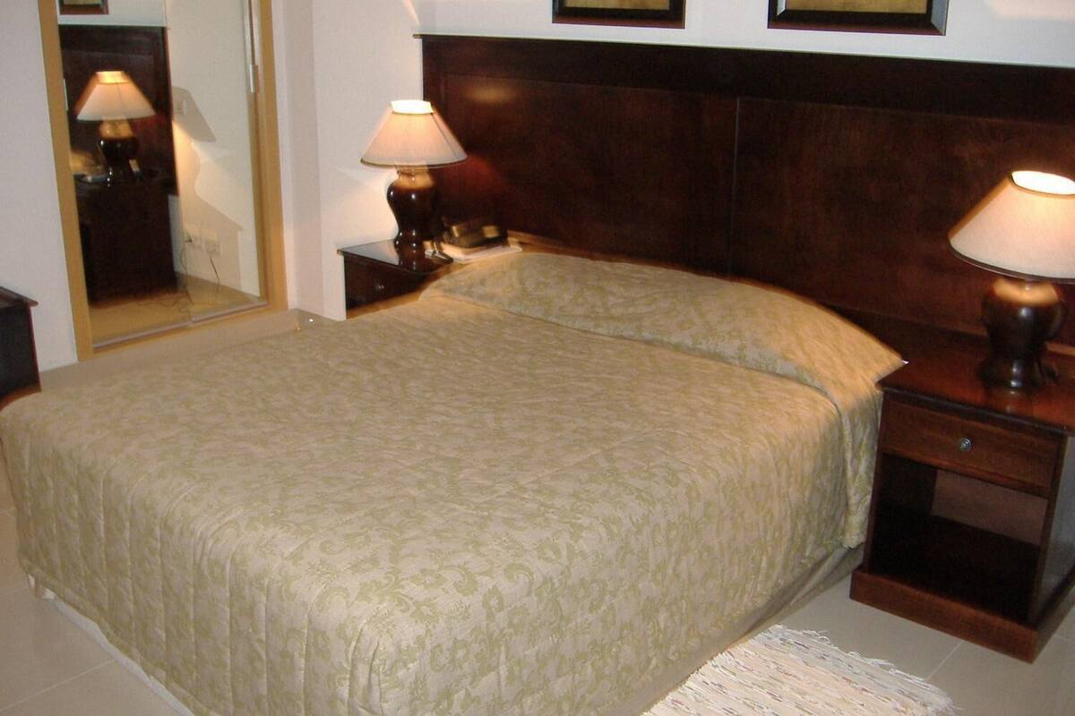 Una cama bien hecha con una colcha estampada, dos lámparas de noche, una mesita de madera y un espejo en la pared.