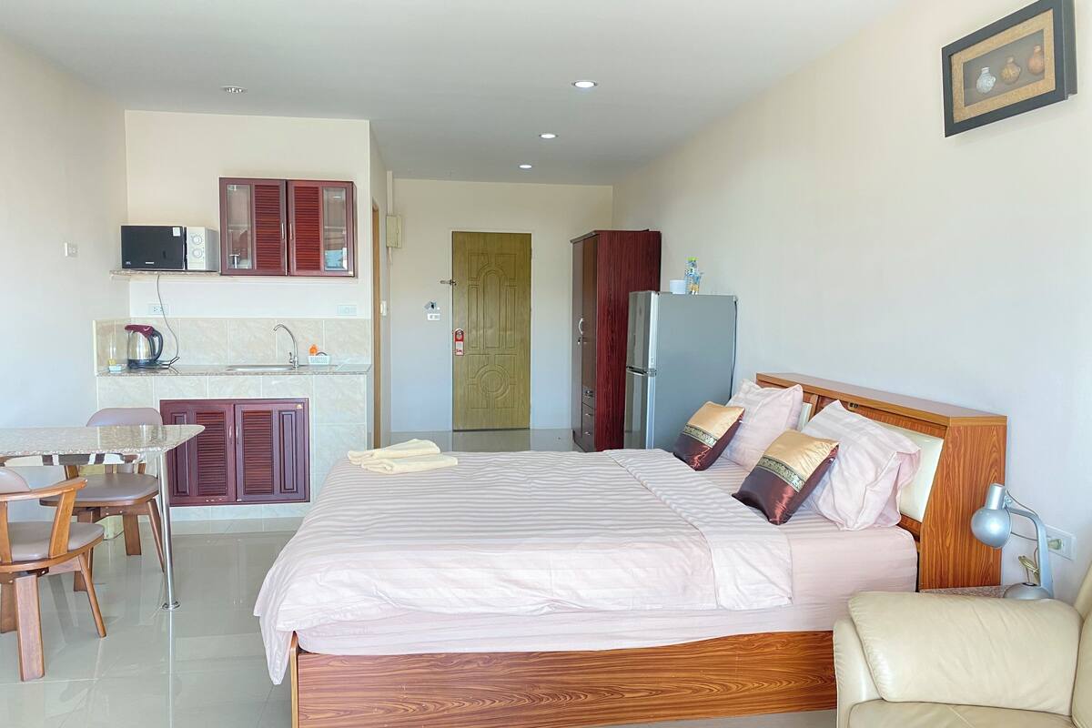 Habitación de hotel con cama, cabecero de madera, escritorio con silla, kitchenette y puerta que da a otra habitación.