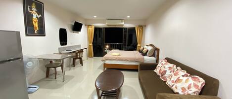 A modern hotel room with a bed, a sofa, a dining table with chairs, a small kitchen area, and a TV mounted on the wall.
