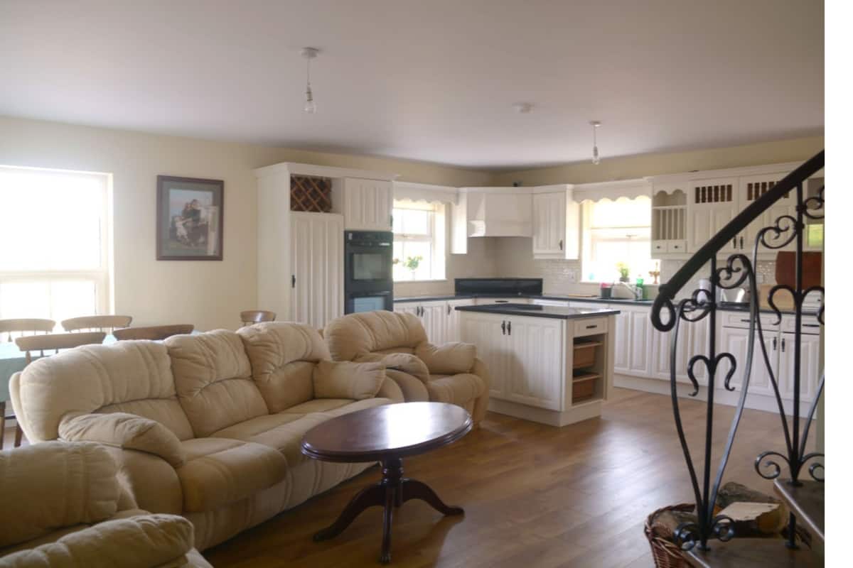 A spacious living room with a beige sectional sofa, a round coffee table, and a kitchen area with white cabinets and a black countertop.