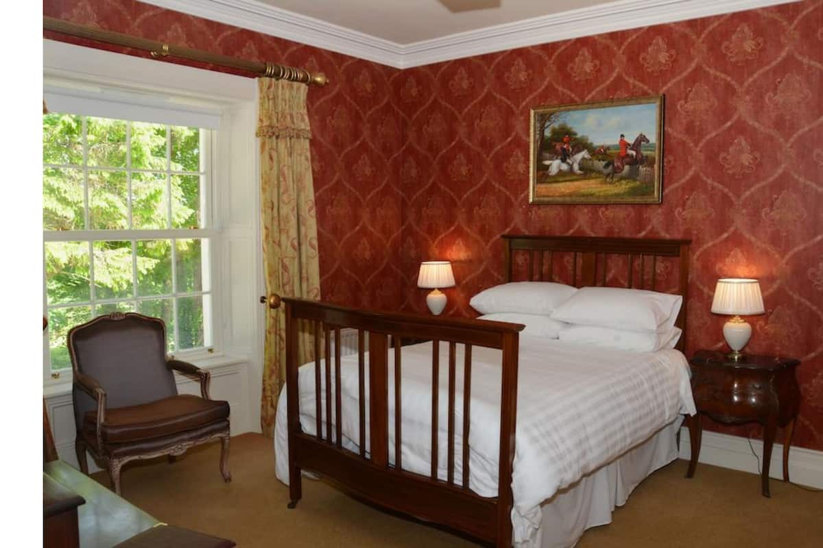 A bedroom with a wooden bed, two bedside lamps, a chair, and a window with curtains.