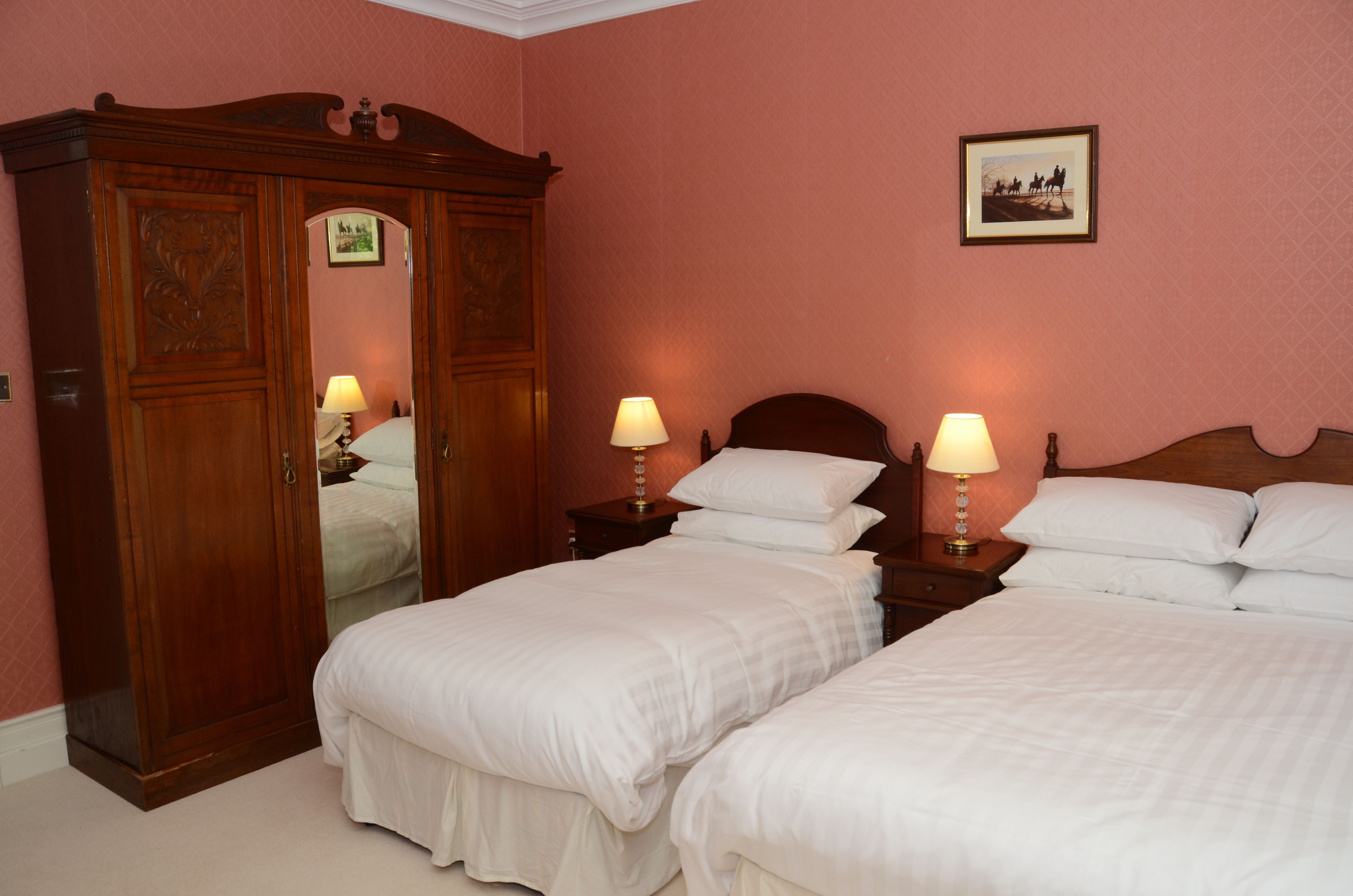 A hotel room with two single beds, a wooden wardrobe, bedside tables with lamps, and a framed picture on the wall.