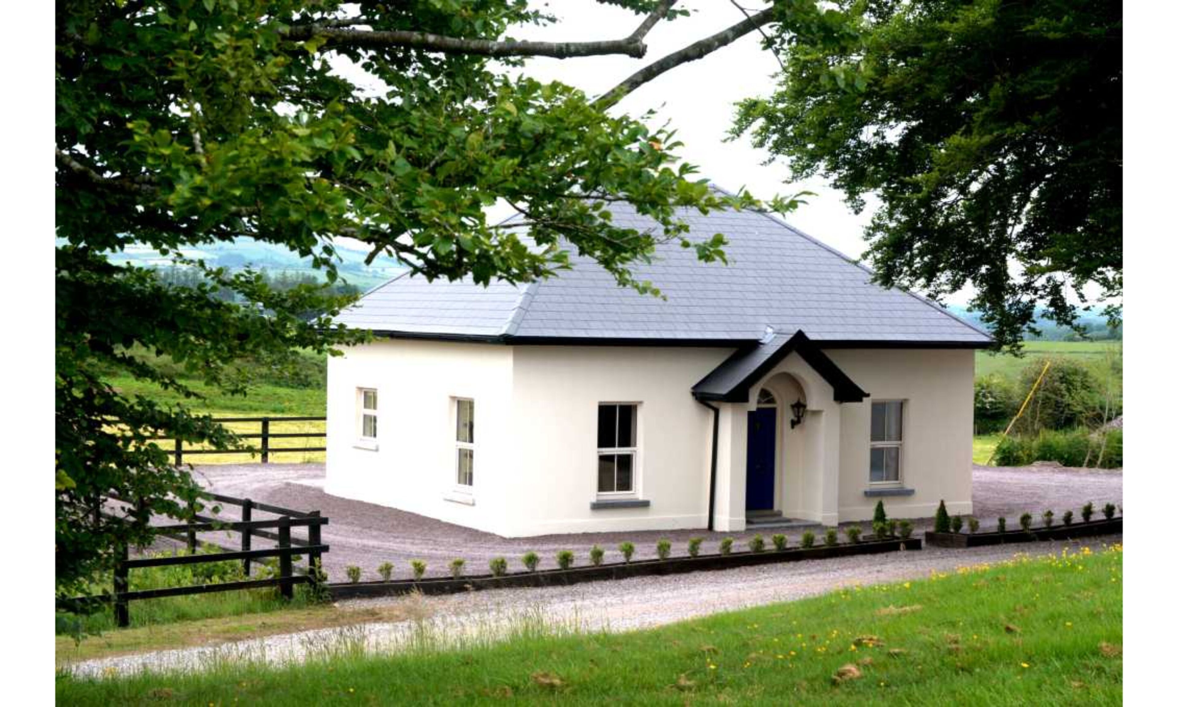 Une petite maison de plain-pied, avec un toit sombre, des murs blancs et une porte bleue. Elle est entourée d’une allée gravillonnée et d’une clôture en bois.