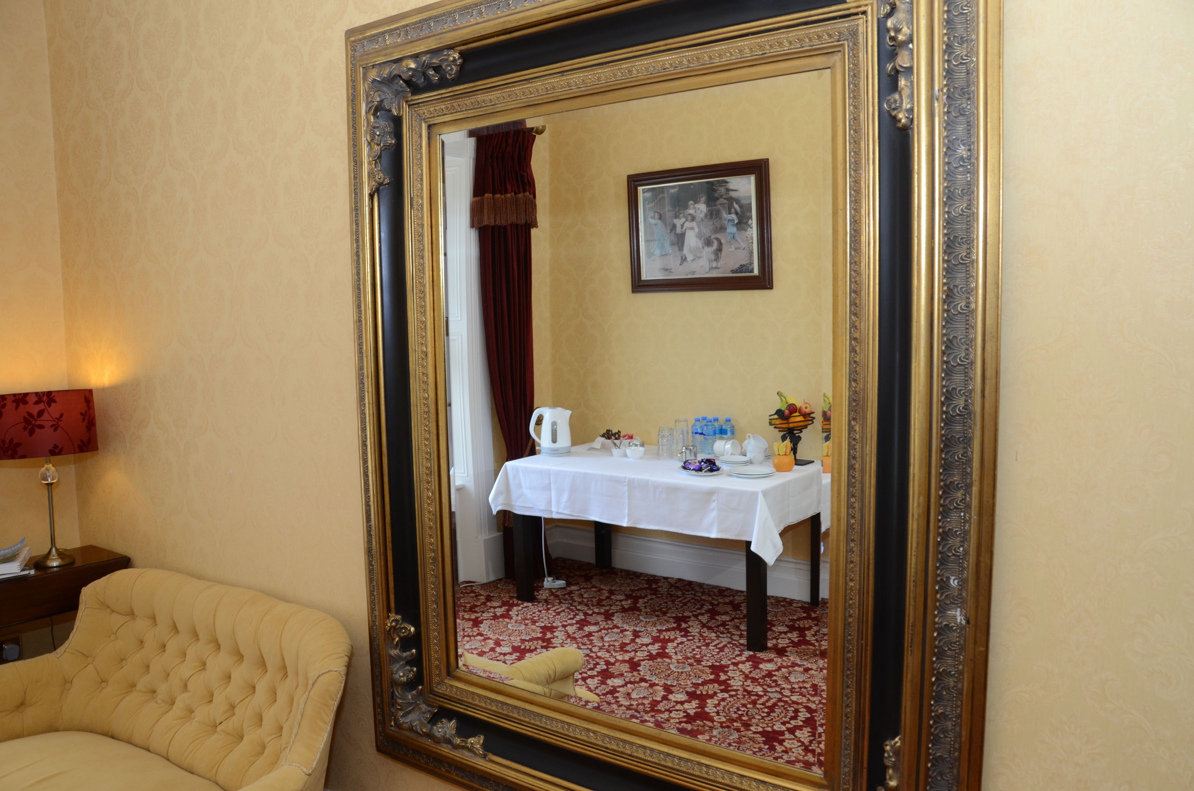 A room with a large ornate mirror, a white table with a red patterned tablecloth, and a beige armchair.