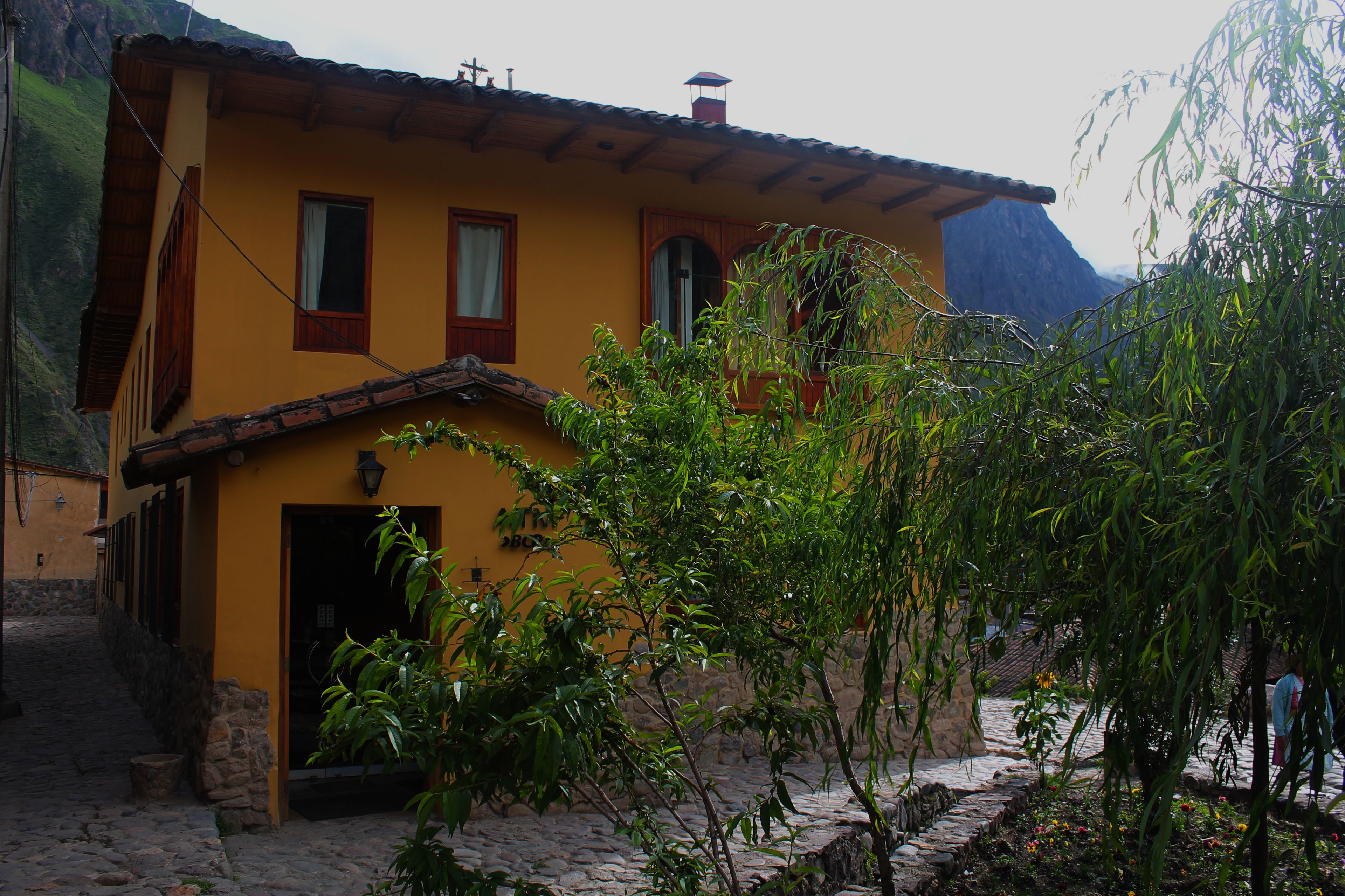 A yellow building with a stone staircase leading to a door, surrounded by greenery and a mountain in the background.