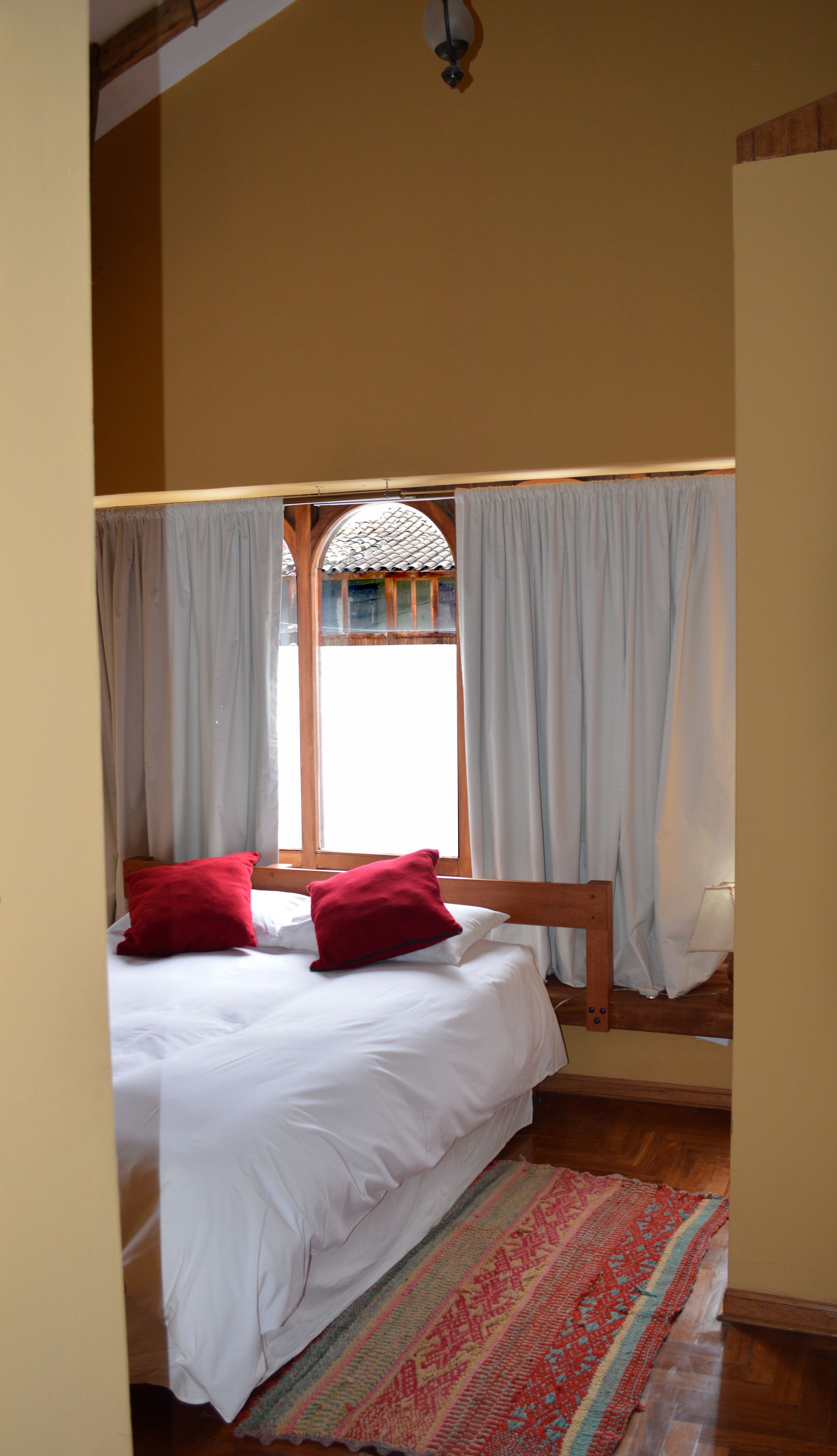 A bedroom with a bed, two red pillows, a wooden headboard, a window with curtains, and a patterned rug.