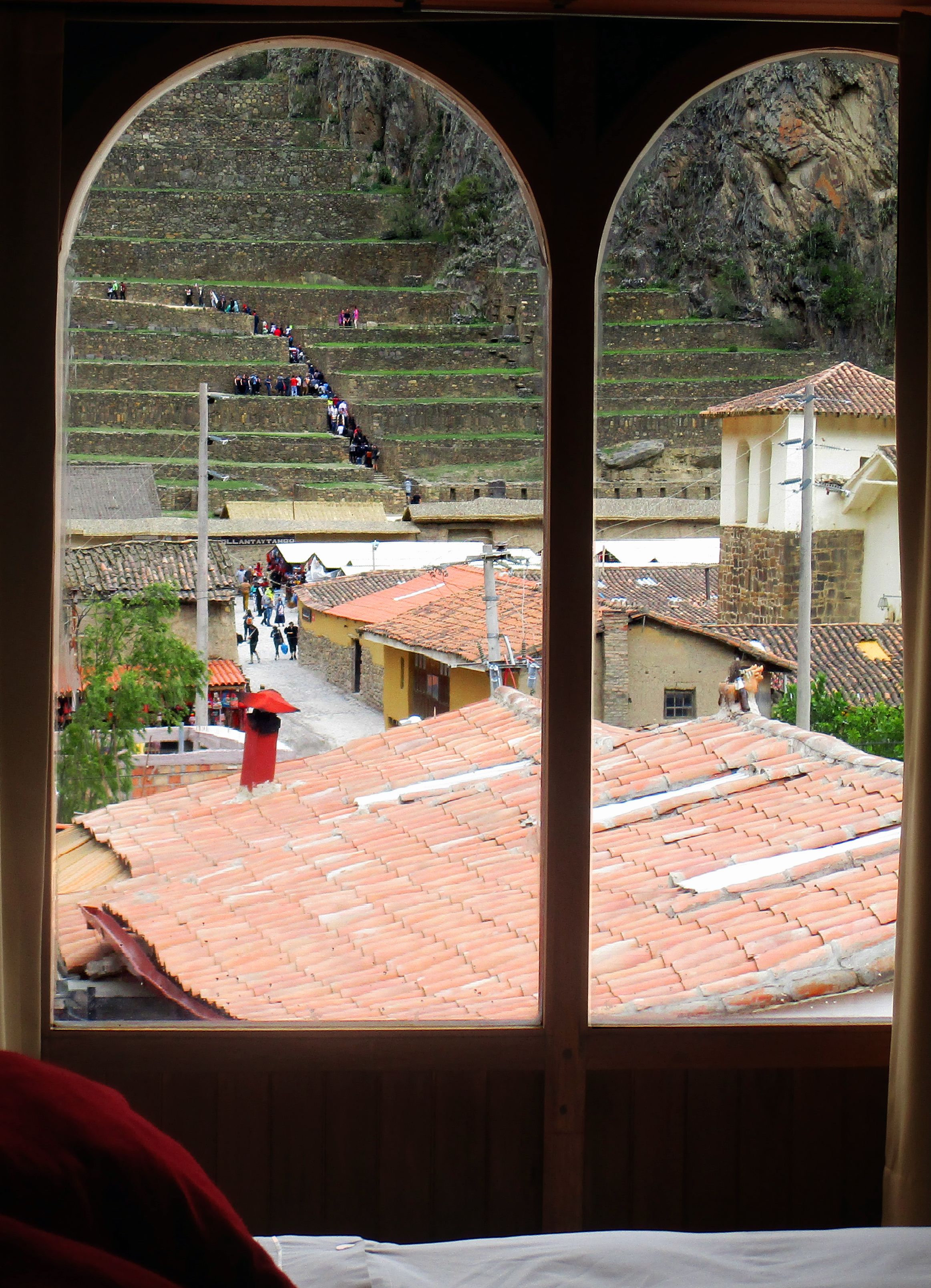 A view through an arched window of a building with terracotta tiled roofs, a staircase leading to a hillside, and a small building with a red roof.