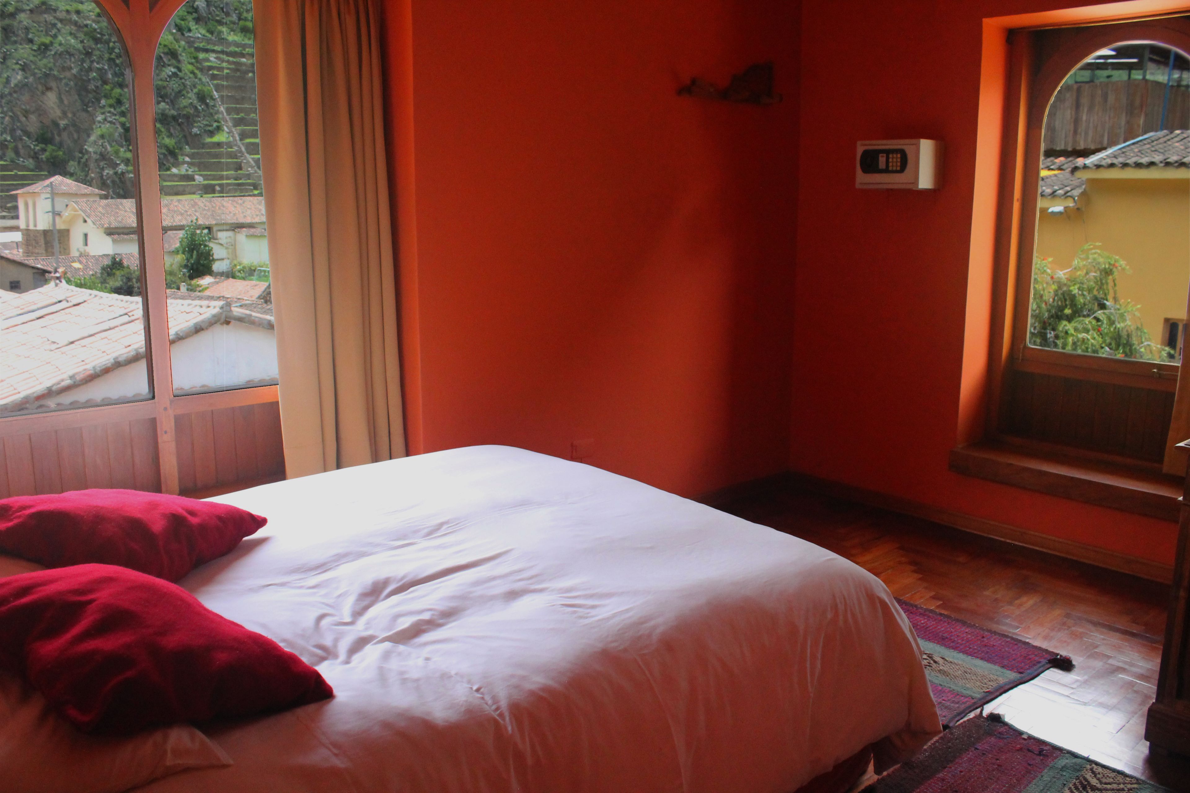 A hotel room with a bed, red pillows, a window with a view of buildings, and a wooden floor.
