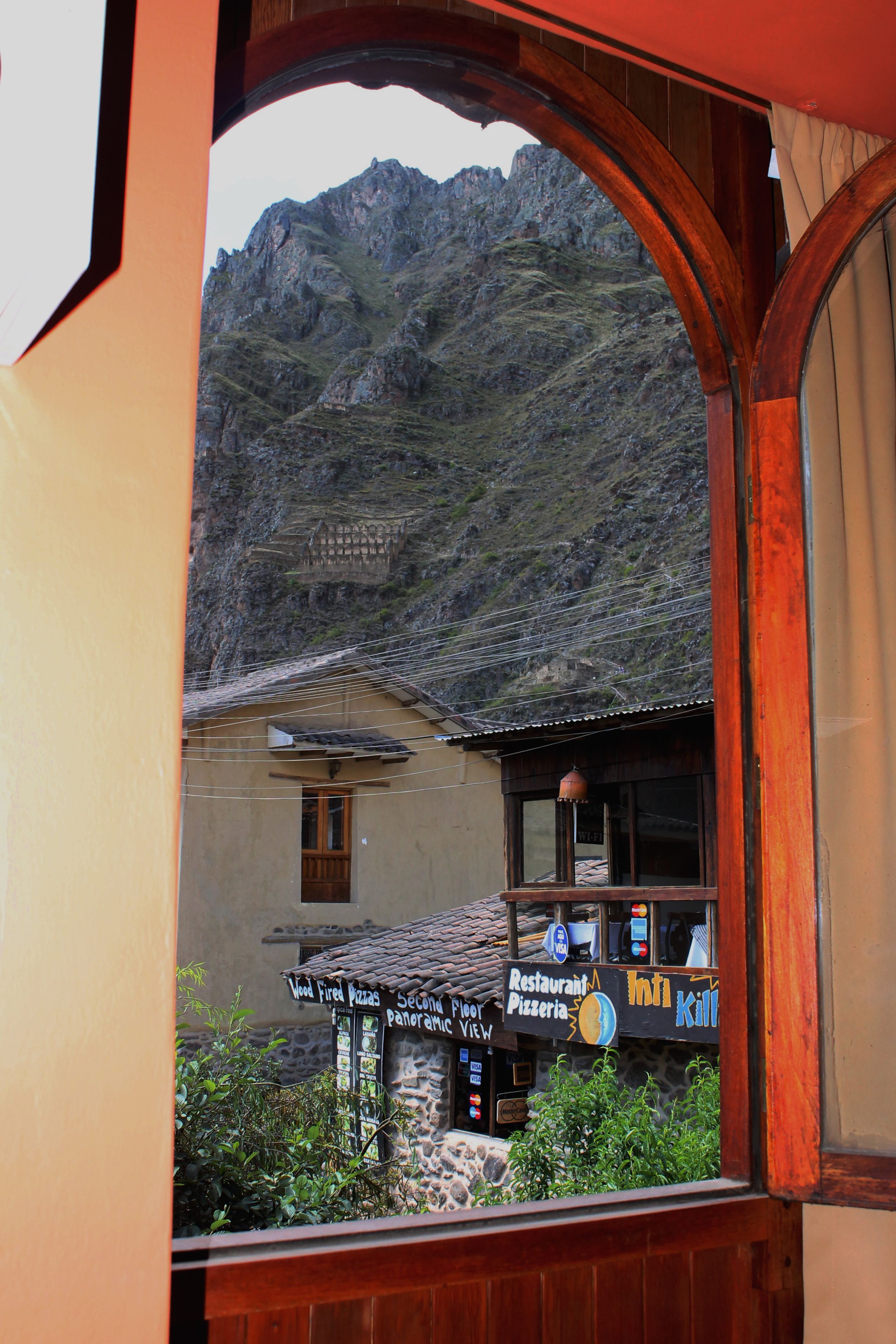 A mountain view through a window with a restaurant sign advertising "First Pizza" and "Second Panoramic View."
