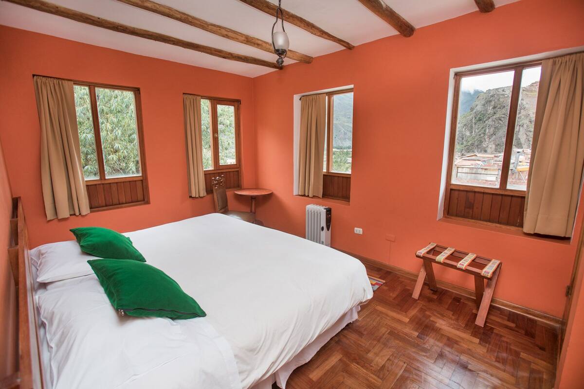 Une chambre avec un grand lit, deux fenêtres avec des rideaux, un radiateur et une petite table en bois.