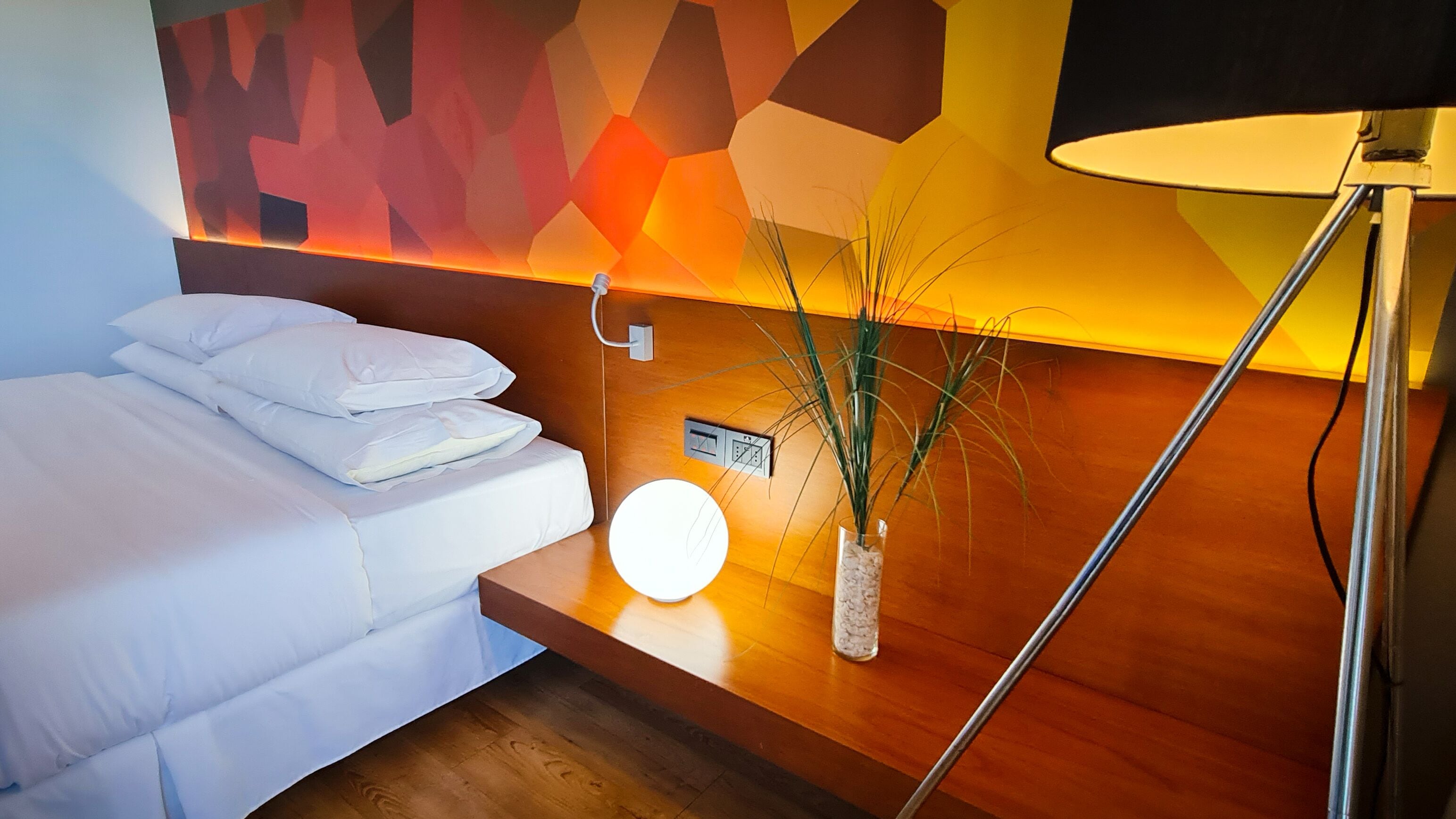 A hotel room with a bed, a bedside table with a lamp, and a wall-mounted control panel.