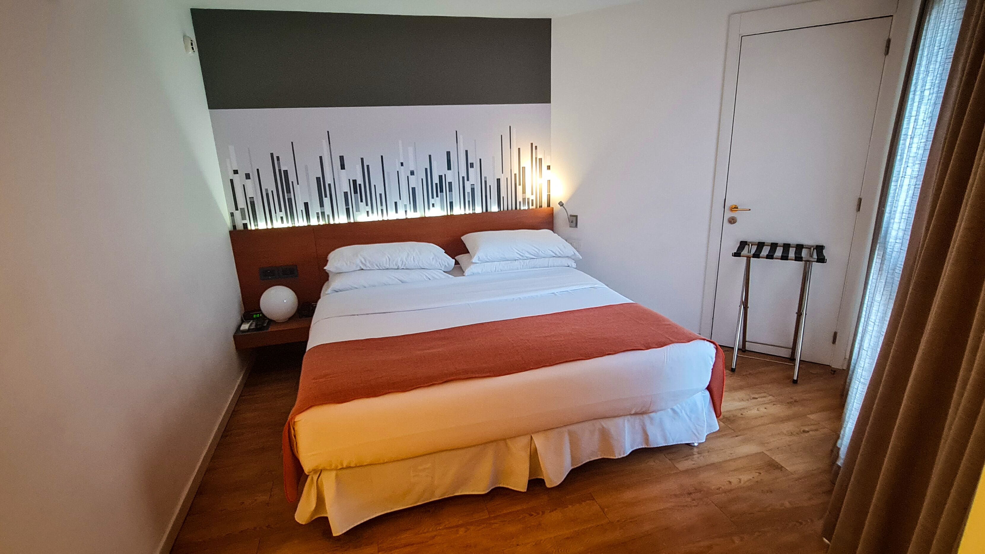 Una habitación de hotel moderna con una cama grande, un cabecero con paneles de luz verticales, una mesita de noche con una lámpara y una puerta con perchero para toallas.
