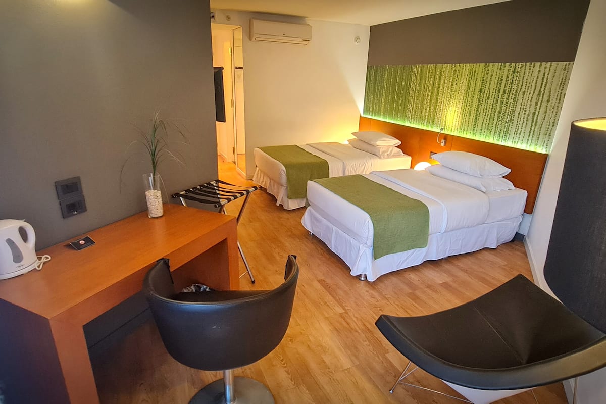 A hotel room with two beds, a desk, a chair, a lamp, and a wall-mounted air conditioner.