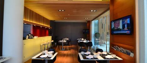 A modern dining area with tables set for service, a bar area, and a television screen displaying a video conference.
