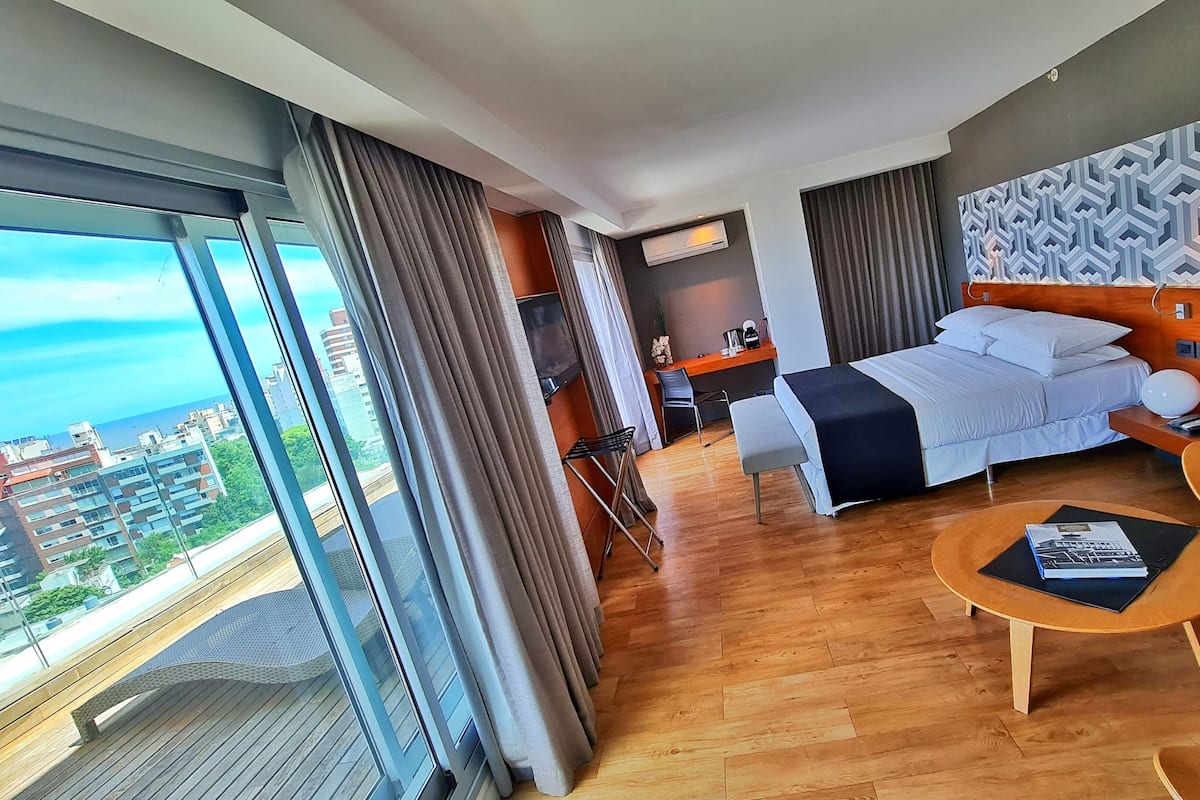 A modern hotel room with a large bed, a desk, a chair, and a view of the city.
