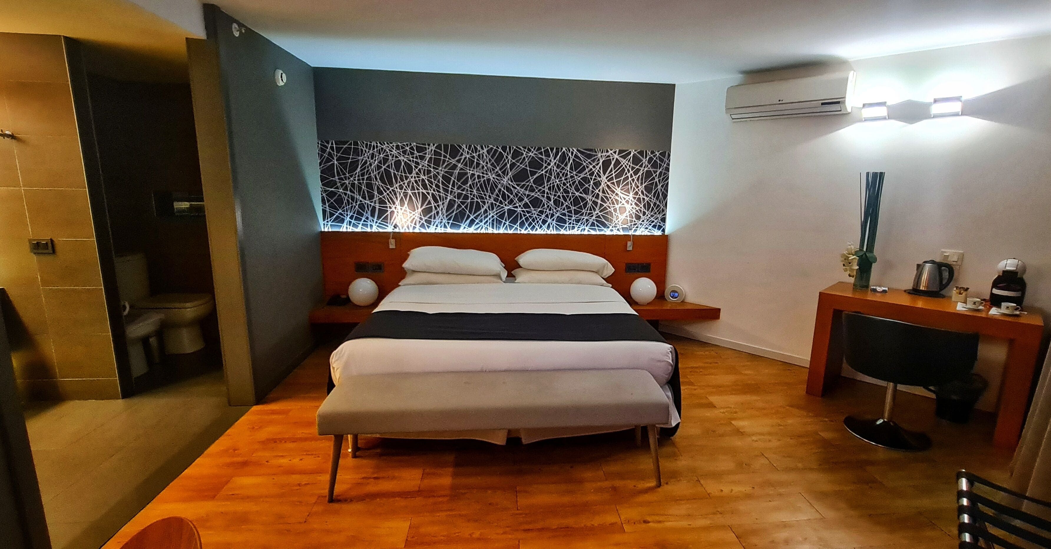 Et moderne hotelværelse med trægulv, en stor seng med hvide sengetøj, et skrivebord med stol og en dekorativ vægpanel.