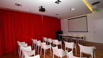 A conference room with a red curtain backdrop, white chairs, a projector screen, and a table with chairs.